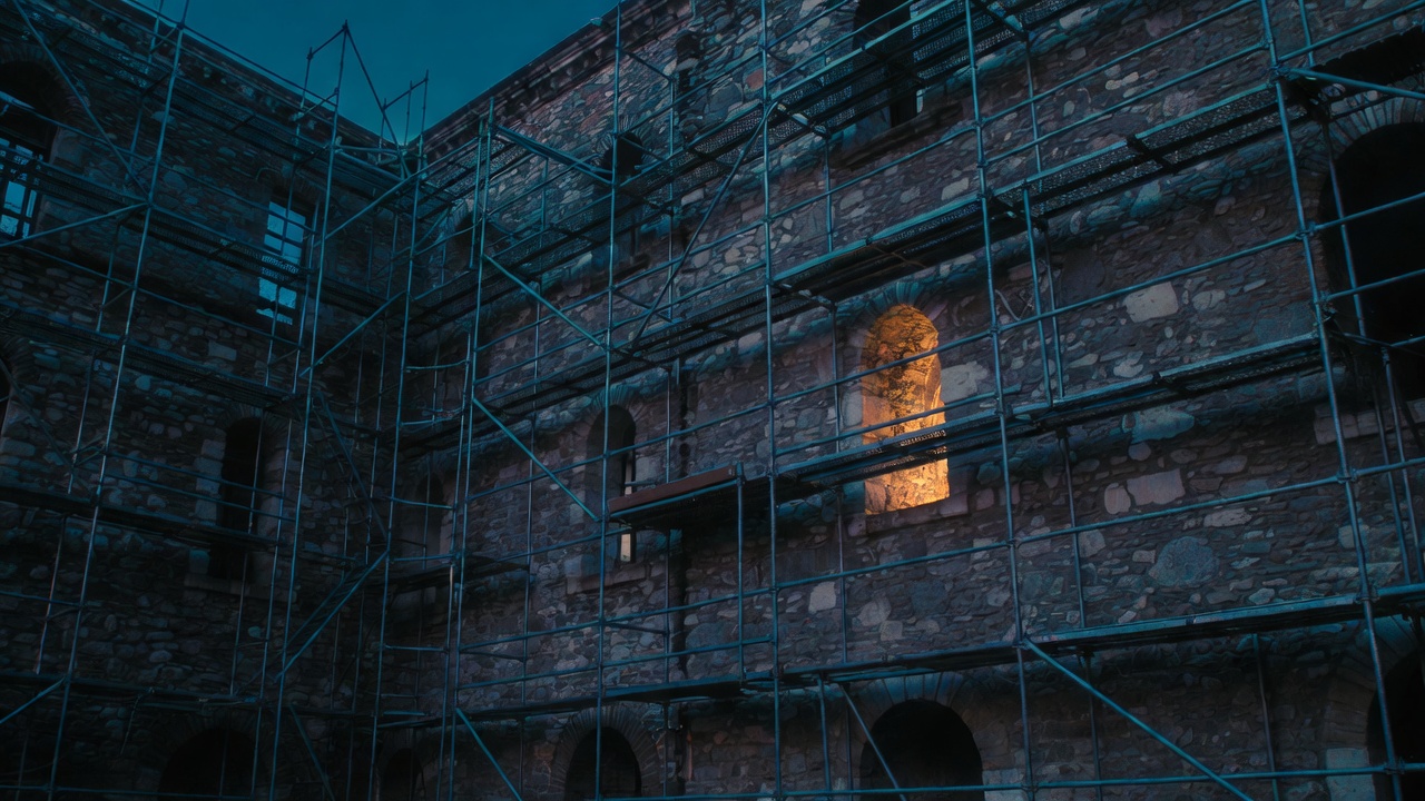 A stone building facade at blue hour partly covered by fine metal scaffolding, one window lit