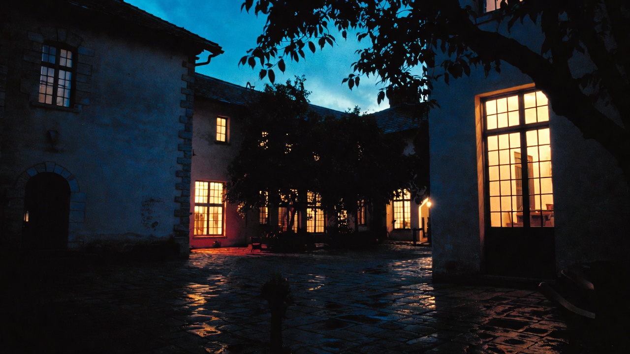 A boutique hotel at dusk, warm interior light spilling through tall windows onto a wet stone terrace